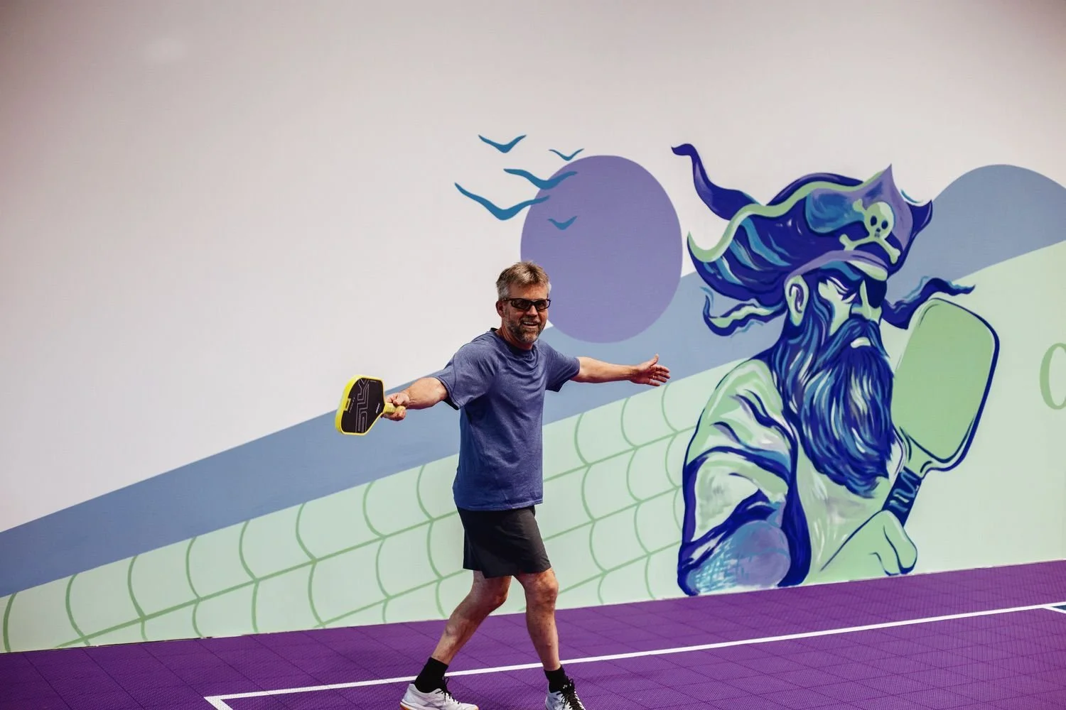 A player on the purple court in front of the wizard mural at Pickleball Cove