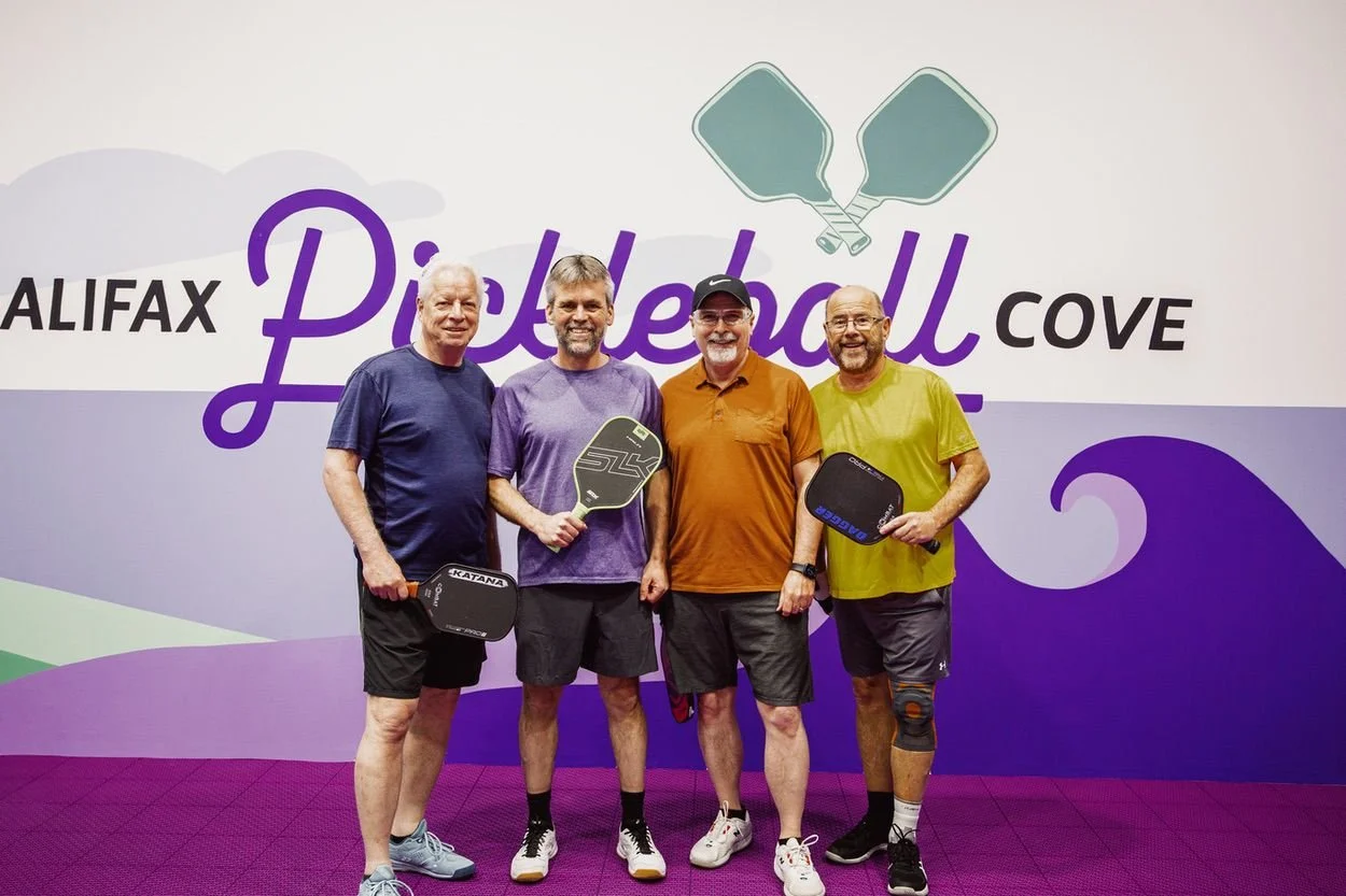 Four players holding paddles in front of the Halifax Pickleball Cove wall sign